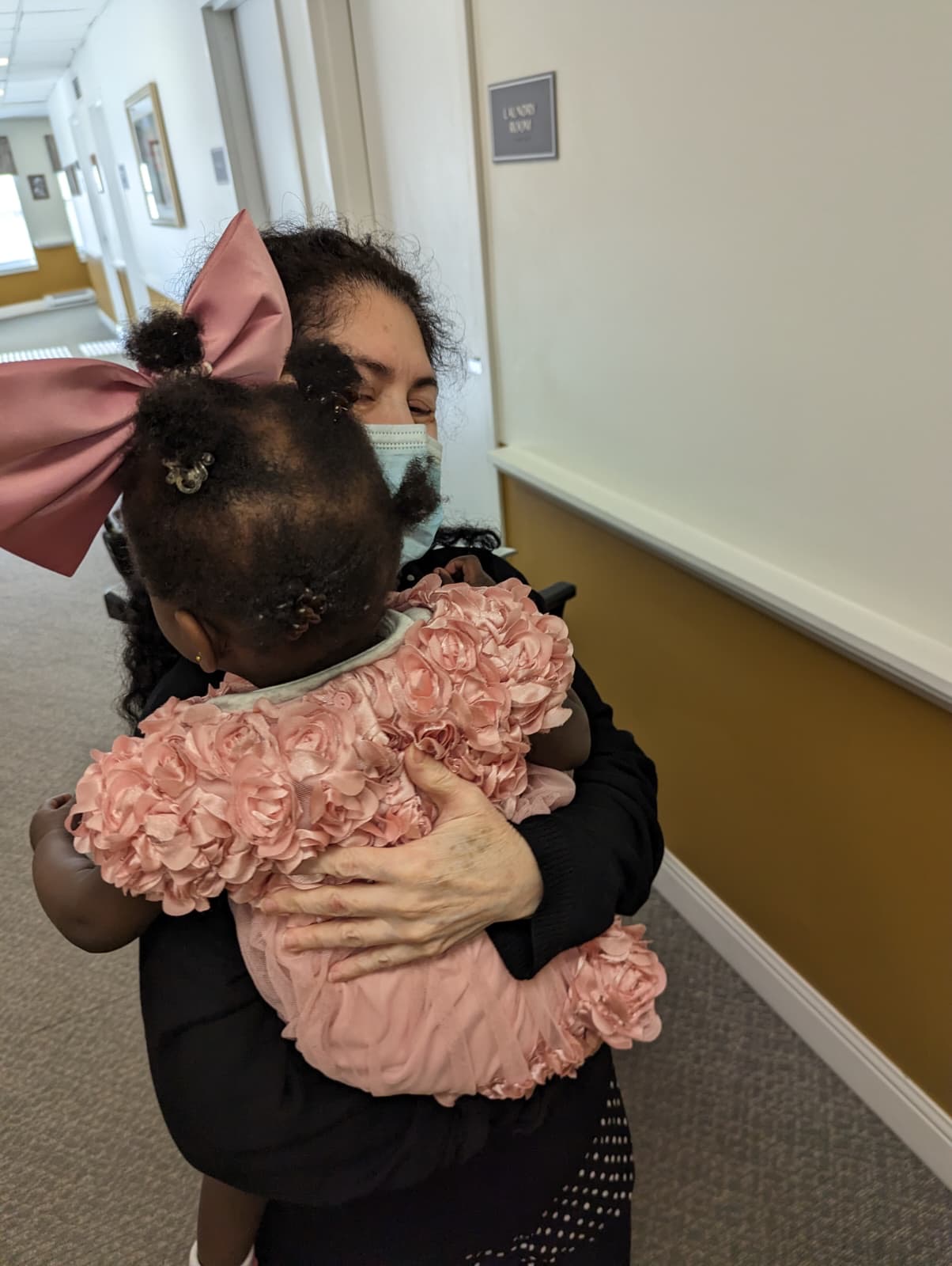 A caregiver in a mask holding a small child in a pink rosette dress in a quiet hallway.