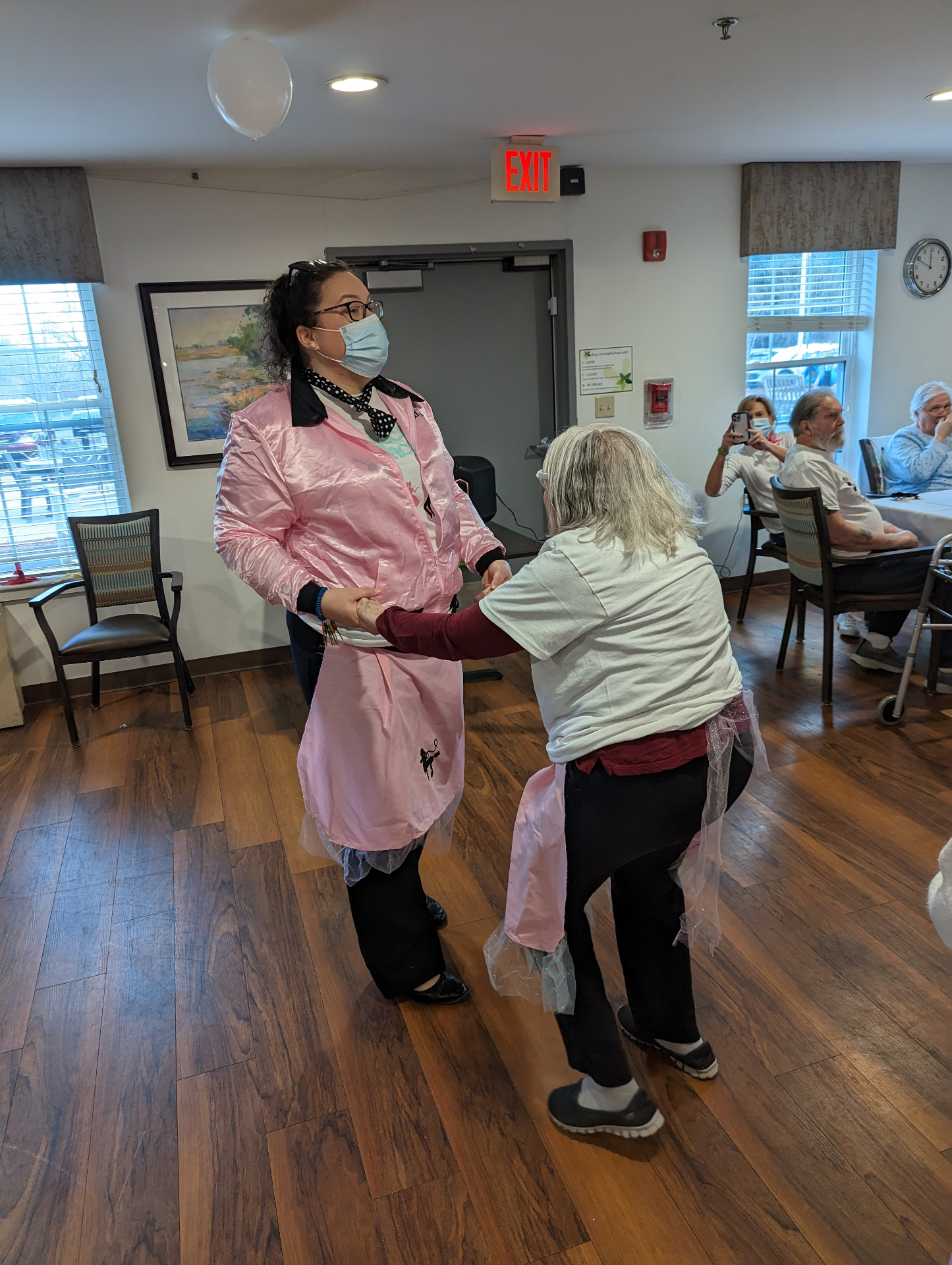 A caregiver in a pink Sock Hop jacket dancing with an older woman on a wood floor.